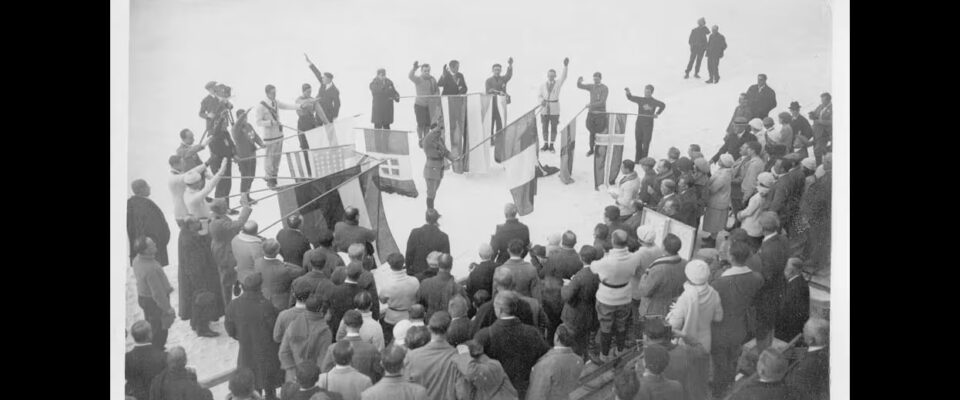 Black and white photo of a crowd of people watching a circle of men holding flags for an Olympic ceremony.
