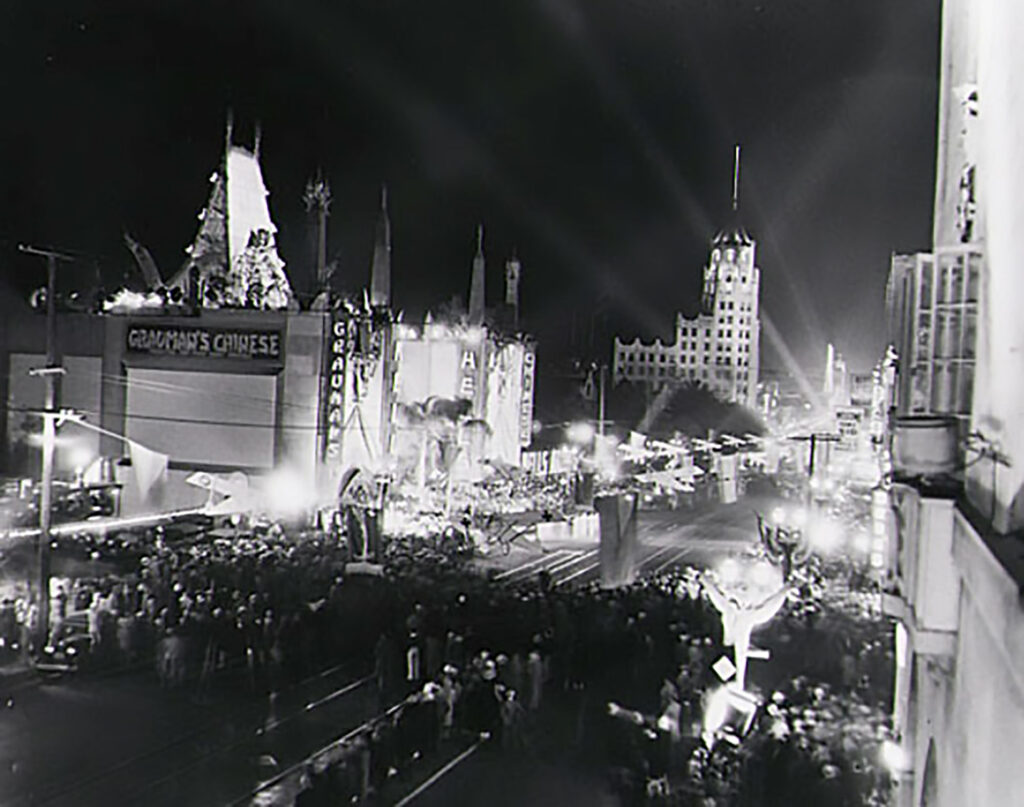 Both sides of a busy urban street are intensely illuminated by lights from a large movie theater and other buildings. A large crowd is gathered in the street.