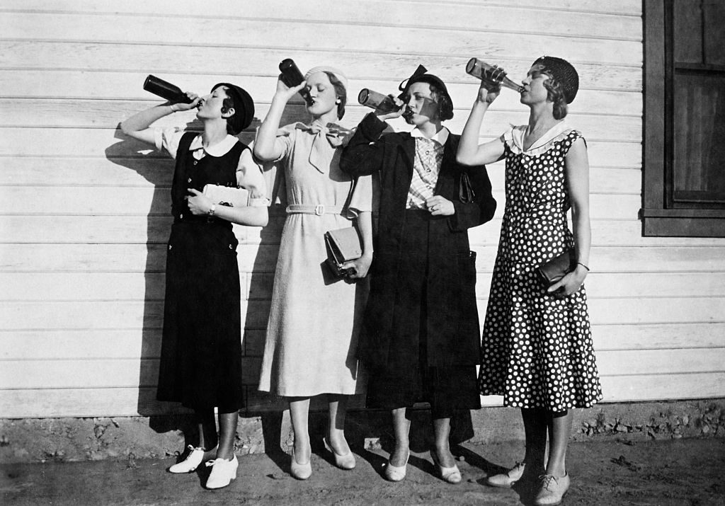 A photo of four white, flapper women lined up along a wall outside, chugging bottles of liquor.