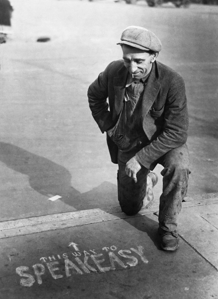 Black and white image of a older man kneeling infront of a side walk were speakeasy was written out on the sidewalk as a form of advertisment.