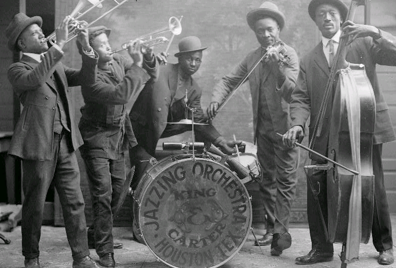 A black and white photograph of a group of five dressed Black men playing in a band. From left to right, they are playing a trombone, a trumpet, the drums, a violin, and an upright bass.