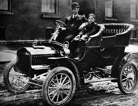 A man and child sit in the front seat of an early automobile. It is stationary on a road.