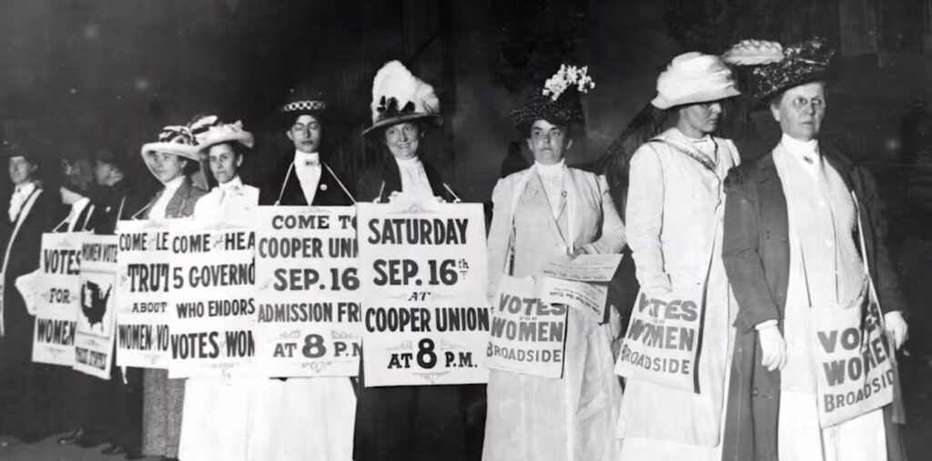 A line of women in formal attire carry signs, wear bags, or hold fliers sharing the date and time of a pro-suffrage event.
