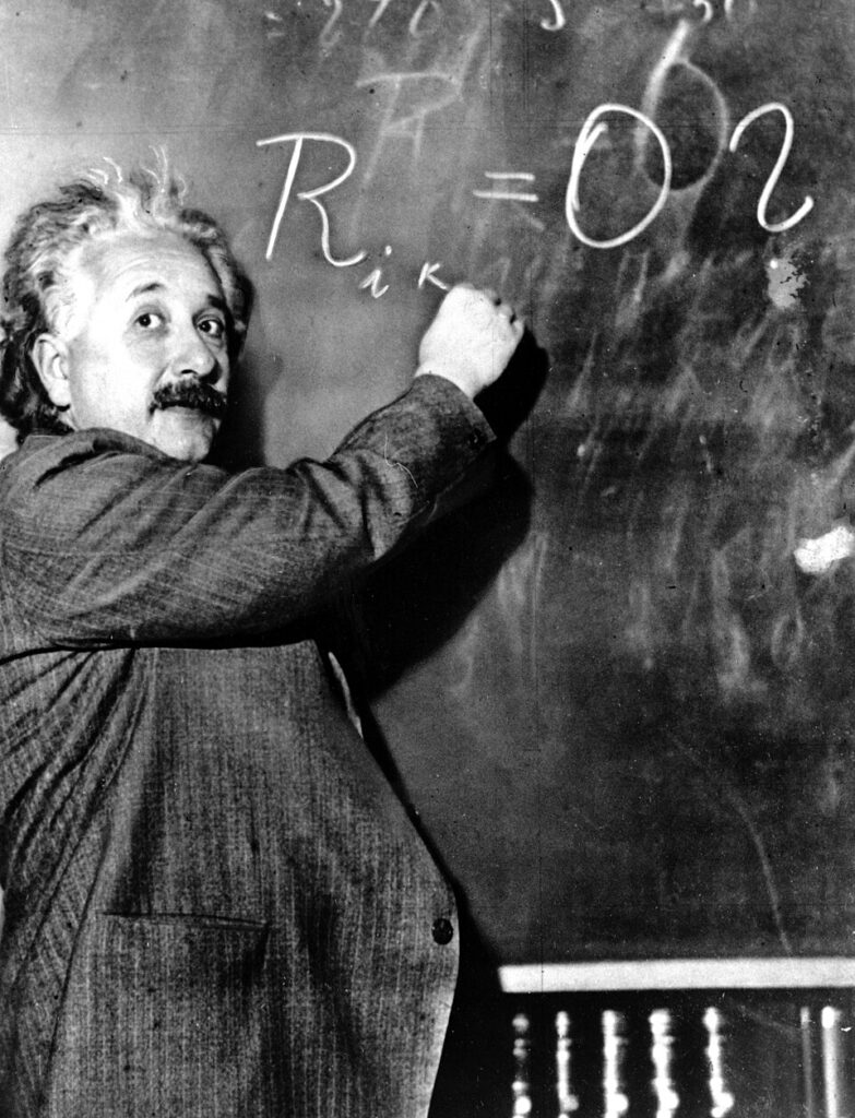 Einstein writes equations on a blackboard while looking back at the camera.