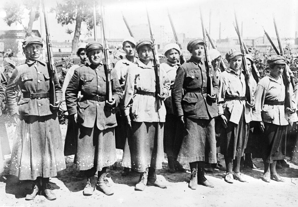 Two rows of women in trench coats are holding gun awaiting orders. The women have their hair short, similar to a man.
