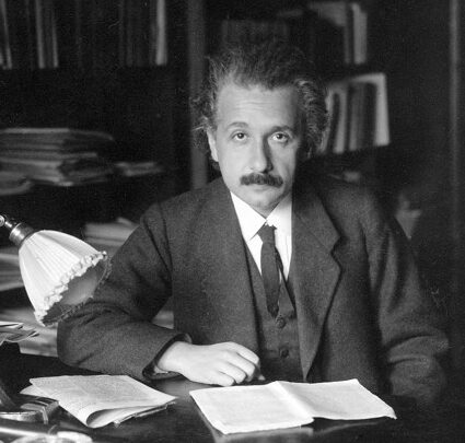 A man sits at a desk with many books around him, and one notebook open in front of him.