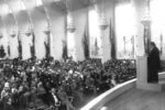 Einstein stands at a podium in front of a large auditorium.