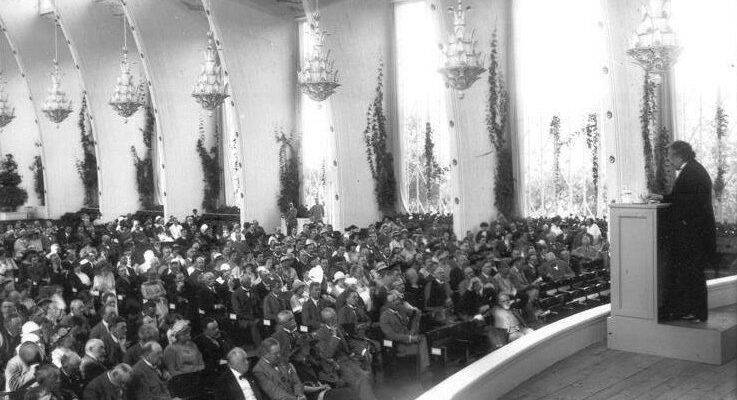 Einstein stands at a podium in front of a large auditorium.