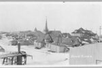A black and white photograph depicting the small town of Nome, Alaska. It is covered in snow. Most of the buildings are very small, with a church steeple rising above the rest of the structures. There are no paved roads pictured in the town. The picture was taken in 1916, less than ten years before the 1925 Serum Run.