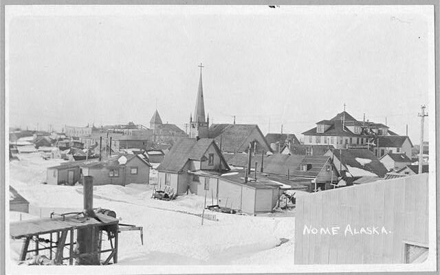 A black and white photograph depicting the small town of Nome, Alaska. It is covered in snow. Most of the buildings are very small, with a church steeple rising above the rest of the structures. There are no paved roads pictured in the town. The picture was taken in 1916, less than ten years before the 1925 Serum Run.