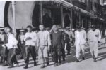 A group of men bound by rope walk down the street with police officers escorting them.