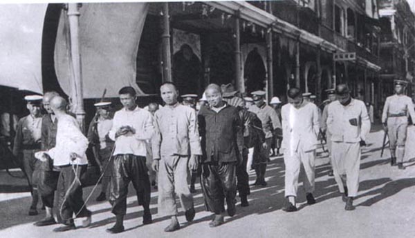 A group of men bound by rope walk down the street with police officers escorting them.