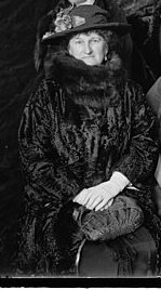 Abby Aldrich Rockefeller is sitting down. She has short curly hair. She is wearing a huge fur coat. She is also wearing a hat with a flower on it.