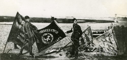 A black and white photo of two men in military uniforms are in a flat plain. Beside the two men are four Soviet flags. All four flags have Cyrillic text on it. One flag is being held by one of the man and it has a hammer and sickle on it.