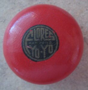 A red wooden yo-yo on a stone surface. There is a graphic on the yo-yo that is a black circle with faded brown text reading "FLORES YO-YO."