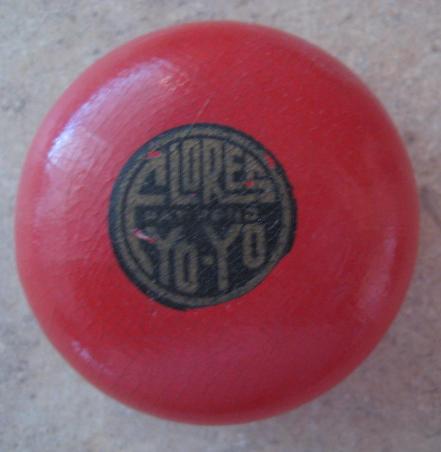 A red wooden yo-yo on a stone surface. There is a graphic on the yo-yo that is a black circle with faded brown text reading "FLORES YO-YO."