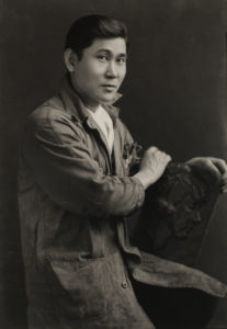 Soichi Sunami is sitting sideways on the chair. His back is not against the back of the chair. He is wearing a trenchcoat.