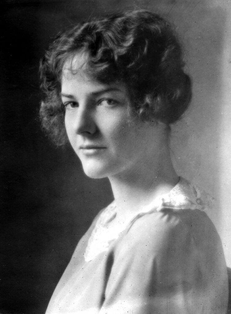 Abby Rockefeller is facing to the side while her head is looking at the camera. She has short hair and wearing a dress.