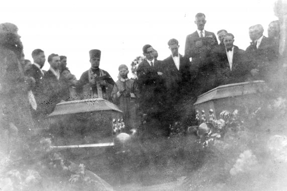 A black and white photograph of a funeral. Two caskets sit in the foreground, and people dressed in black stand behind them.