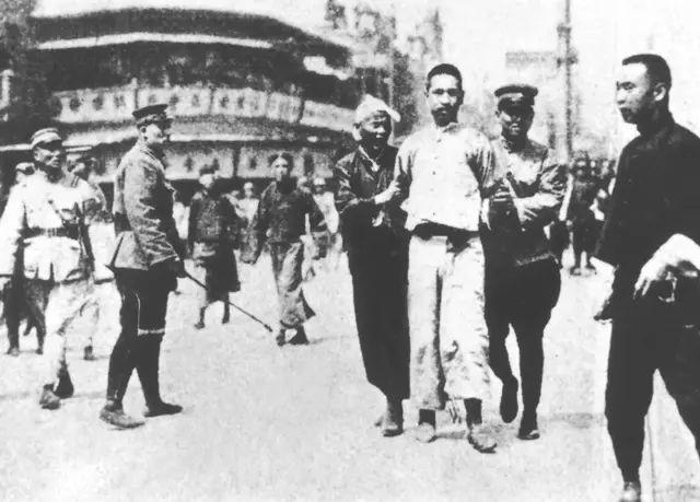 A prisoner is escorted through the streets of a city by guards.